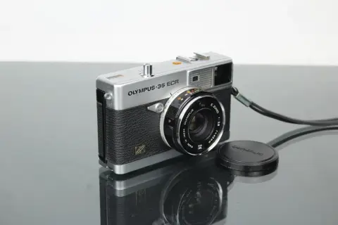 A vintage silver Olympus 35 ECR rangefinder camera with a black textured grip sits on a reflective surface.