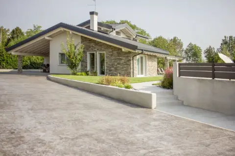Un moderno **pavimento in cemento per esterno** accoglie una casa con tetto spiovente e rivestimento in pietra.