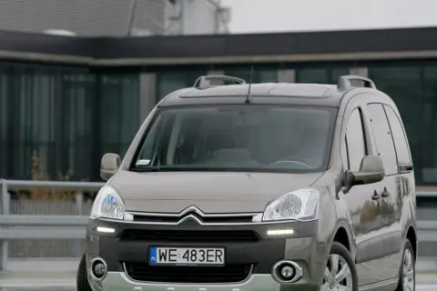 Szary Citroen Berlingo 1.6 HDI z relingami dachowymi zaparkowany przed nowoczesnym budynkiem.