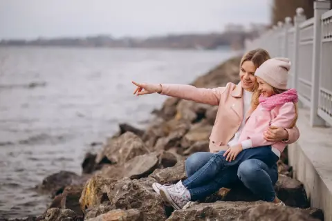 Mama z córką siedzą na kamieniach nad jeziorem. Mama wskazuje coś palcem, a dziewczynka się śmieje. To świetne gąski atrakcje dla dzieci!