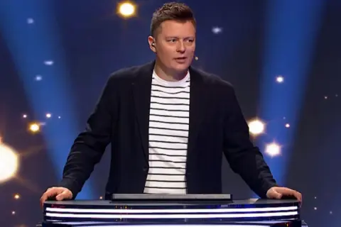 Prowadzący program "Jaka to melodia?" w eleganckim stroju, zastanawia się, ile zarabia zawodowy gracz w Jaka to melodia za jeden odcinek.