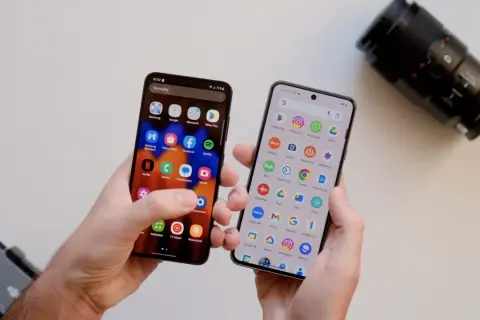 Dwa telefony w dłoniach, jeden z ikoną aparatu, drugi z ikoną aparatu. Który telefon ma lepszy aparat? Obok leży obiektyw aparatu.