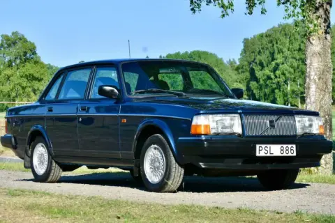 Mörkblå Volvo 240 från 1993 parkerad på en grusväg med grönskande träd och fält i bakgrunden.