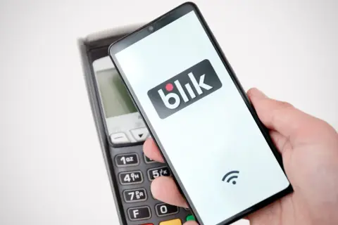 Dłoń trzyma smartfon z logo Blik nad terminalem płatniczym. Szybki przelew na telefon bez obaw o limit.