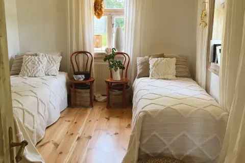Sypialnia w stylu cottage core z dwoma łóżkami, drewnianą podłogą i oknem z firankami.