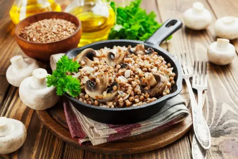 Pyszne kaszotto z pieczarkami w żeliwnej patelni, udekorowane natką pietruszki. Obok świeże pieczarki i miseczka kaszy gryczanej.