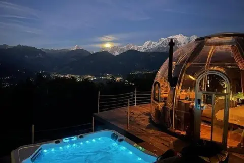 Un jacuzzi illuminé et une bulle transparente, un logement insolite montagne offrant une vue spectaculaire sur les sommets enneigés et la vallée scintillante.