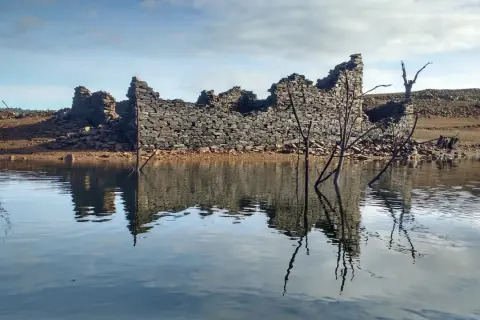 Ruinas de piedra emergen del agua en el embalse de Portodemouros, reflejando su silueta en la calma superficie.