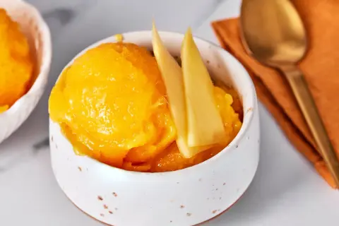 Erfrischendes Mango-Sorbet in einer Schale, garniert mit Mangostücken. Daneben liegt ein goldener Löffel auf einem orangen Tuch.