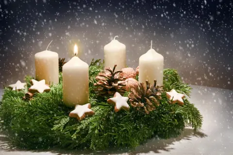 Ein Adventskranz mit vier Kerzen, Tannenzweigen, Zapfen und Sternen. Die erste Kerze brennt. Der Ursprung des Adventskranzes liegt im 19. Jahrhundert.
