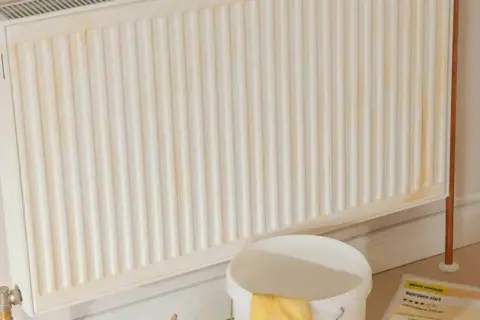 Ein weißer Heizkörper mit gelblichen Flecken, bereit zum heizung streichen. Daneben ein Eimer mit gelben Handschuhen und Malerzubehör.