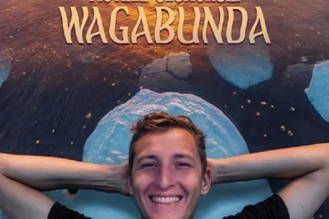 Uśmiechnięty Piotr Szumowski, gwiazda stand-upu, leży na tle napisu "WAGABUNDA" i lodowych krajobrazów.