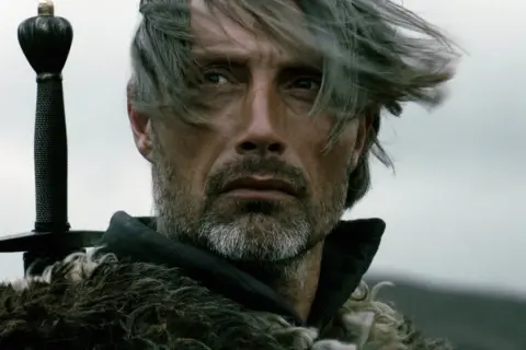 Mads Mikkelsen mit Schwert und Pelzmantel, ein Bild, das an seine Rollen in Filmen und Serien erinnert.
