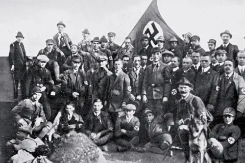 Grupa mężczyzn w mundurach i kapeluszach, z flagą NSDAP w tle. To zdjęcie pokazuje członków partii nazistowskiej, czyli NSDAP.