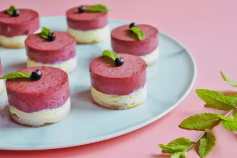 Urocze mini serniczki z jagodami i miętą na jasnoniebieskim talerzu, gotowe do degustacji.