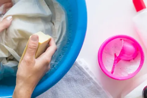 Hände reiben mit Kernseife über ein Kleidungsstück in einer blauen Schüssel, um fettflecken aus kleidung entfernen. Waschmittel und Flaschen daneben.