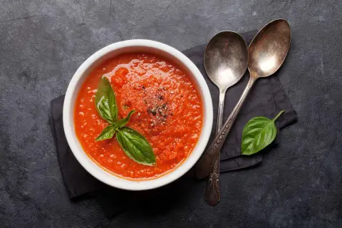 Schüssel mit bester Tomatensauce, garniert mit Basilikum und Pfeffer, daneben zwei Löffel auf dunklem Hintergrund.