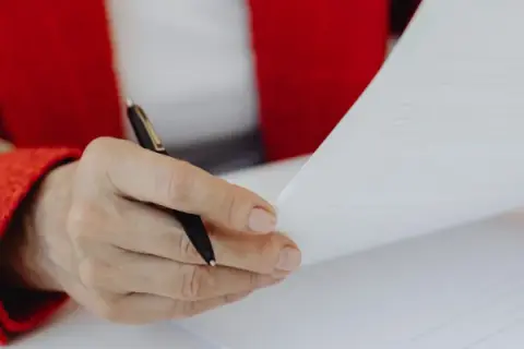 Dłoń w czerwonej marynarce trzyma długopis nad dokumentem, zastanawiając się, czy umowa zlecenie wlicza się do emerytury.