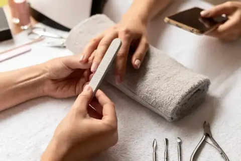 Dłonie przygotowują się do zabiegu, jak zdjąć paznokcie żelowe. Pilniczek delikatnie modeluje paznokieć, a obok leżą narzędzia do manicure.