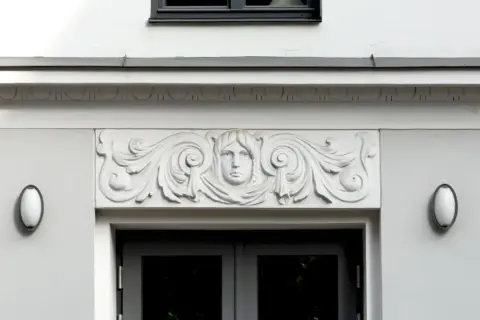 Detalle arquitectónico: una cornisa con un rostro femenino y volutas decorativas sobre una puerta.