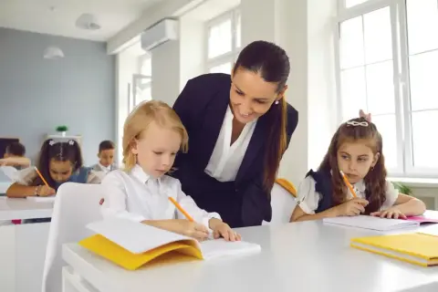 Nauczycielka podczas hospitacji wspiera ucznia w pisaniu. Dzieci siedzą przy ławkach, skupione na zadaniu.
