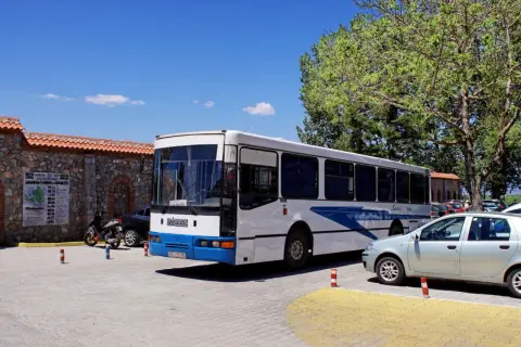 Biały autobus z niebieskimi pasami, oznaczony jako "Arbus", stoi zaparkowany obok kamiennego muru. To autobus z Macedonii, gotowy na kolejną podróż.