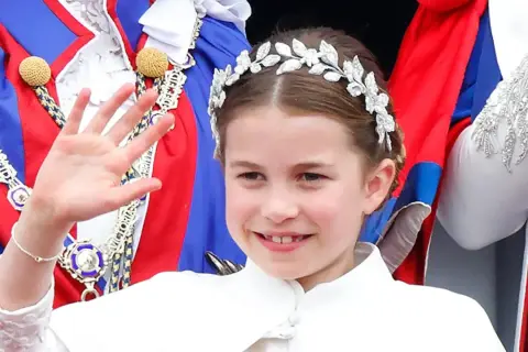 Uśmiechnięta księżniczka Charlotte w tiarze macha ręką, ubrana w białą sukienkę i ozdobne szaty.
