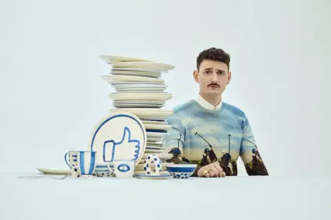 Mężczyzna z wąsikiem w swetrze z motywem polo, obok stosu talerzy i zastawy z symbolem "like". Stylizacja jak z lat dwudziestych.