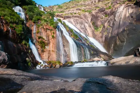 La cascada Fervenza se despliega en múltiples hilos blancos sobre rocas rojizas y verdes, desembocando en un lago sereno.