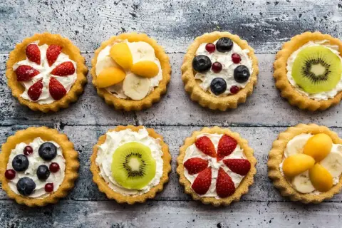 Osiem kolorowych tartaletek z owocami: truskawkami, borówkami, kiwi, bananami i morelami na kremowym nadzieniu.