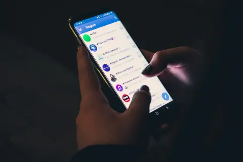 Dłonie trzymają smartfon z otwartą aplikacją Telegram. Czy telegram jest bezpieczny? Aplikacja wyświetla listę czatów i kanałów.