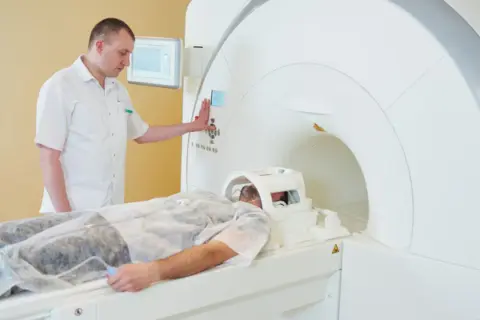 Pacjent leży w aparacie do rezonansu magnetycznego. Technik nadzoruje badanie, przygotowując rejestrację.