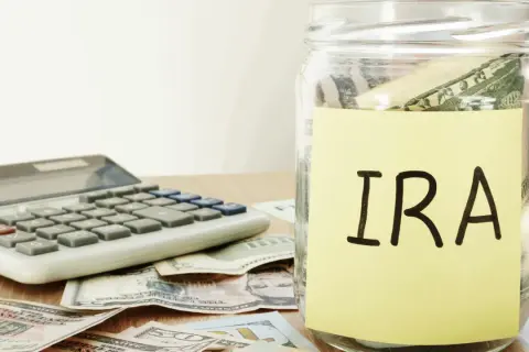 A glass jar labeled "IRA" sits on a table with US dollar bills and a calculator, symbolizing financial planning and the potential for an inherited IRA.