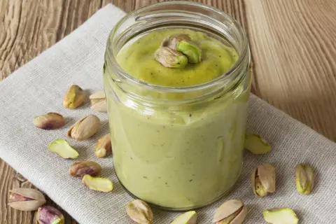 Tarro de crema pistacho casera, decorado con pistachos enteros y trozos, rodeado de más pistachos sobre tela y madera.