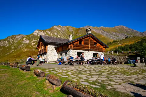 Un rifugio di montagna in pietra e legno, con persone sedute ai tavoli all'aperto, circondato da prati verdi e montagne imponenti sotto un cielo azzurro.