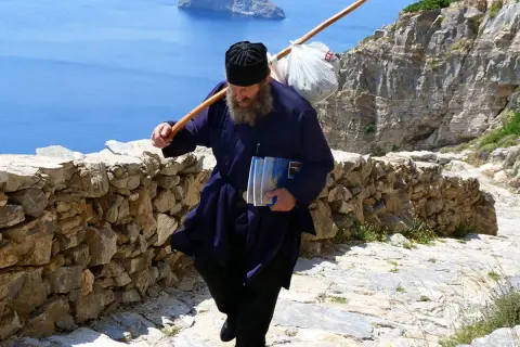 En munk vandrar uppför en stenstig på Amorgos, Grekland, med en stav över axeln och böcker i handen.
