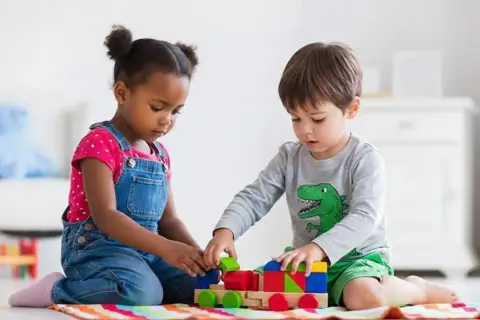 Two children engage in parallel play, building a colorful wooden train with blocks.