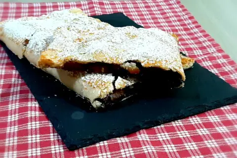 Strudel mele e cioccolato cosparso di zucchero a velo, su una pietra nera e tovaglia a quadri rossi.