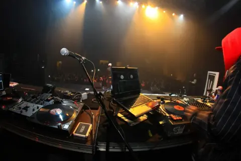 DJ-Pult mit Plattenspielern, Laptop und Mikrofon. Im Hintergrund eine Bühne mit Scheinwerfern und Publikum. Die DJ Namen sind hier nicht sichtbar.