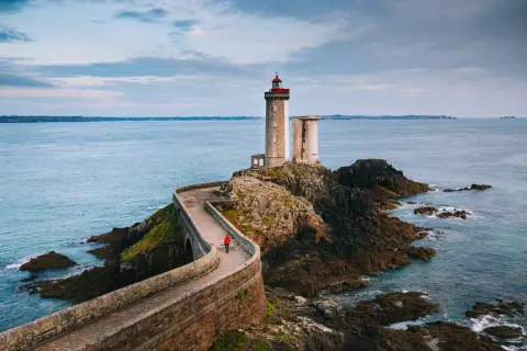 Une personne en manteau rouge marche sur la route des phares, un chemin sinueux menant à un phare sur une île rocheuse.