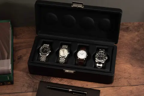 Una caja de reloj negra abierta muestra cuatro relojes de lujo. A la izquierda, libros de Rolex y un bolígrafo.