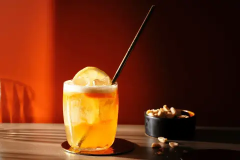 Ein erfrischender Amaretto Sour mit Zitrone und Eiswürfeln, daneben eine Schale Erdnüsse.