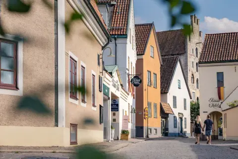 Ett par promenerar hand i hand på en kullerstensgata i Visby, förbi charmiga hus och skyltar som indikerar B&B Gotland och Strand Hotel.