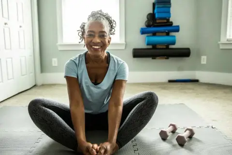 Femme souriante en position de papillon sur un tapis de yoga, prête pour son **gym pour seniors 80 ans**. Haltères roses à proximité.
