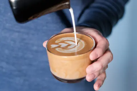 En hand häller mjölk i en kopp för att skapa ett vackert mönster på en flat white.