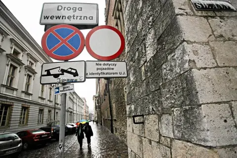 Znaki drogowe informują o zakazie zatrzymywania się i postoju na drodze wewnętrznej. W tle widać ulicę z samochodami i przechodniów.