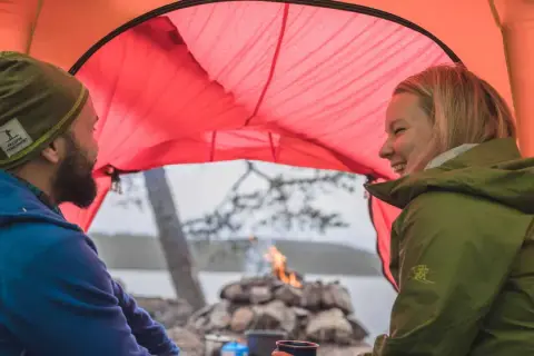 Ett par skrattar inne i ett orange tält. Utanför syns en sjö, träd och en lägereld. Perfekt för att campa i skogen.