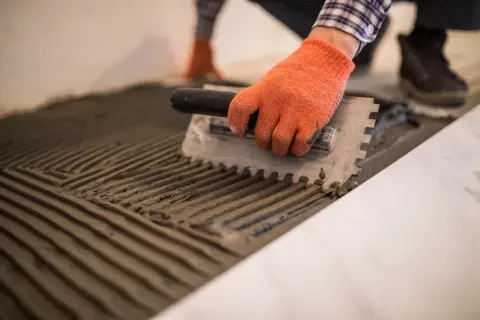 Man con guantes naranjas esparce mortero con llana dentada, preparando el suelo para colocar baldosas.