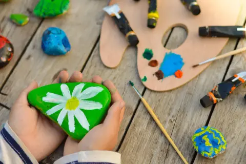 Kreative Ideen zum Steine bemalen für Kinder: Ein grüner Stein mit Gänseblümchen, bunte Farben und Pinsel laden zum Malen ein.
