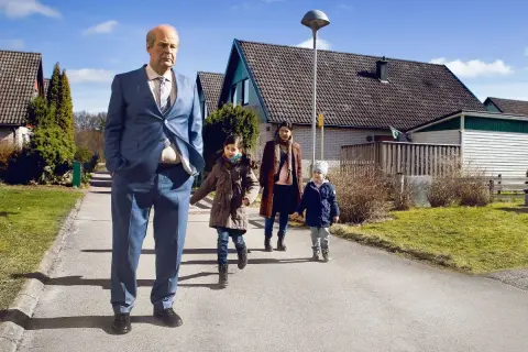 En man i kostym går med en familj i ett villaområde, en scen som kan passa i svenska komedier.